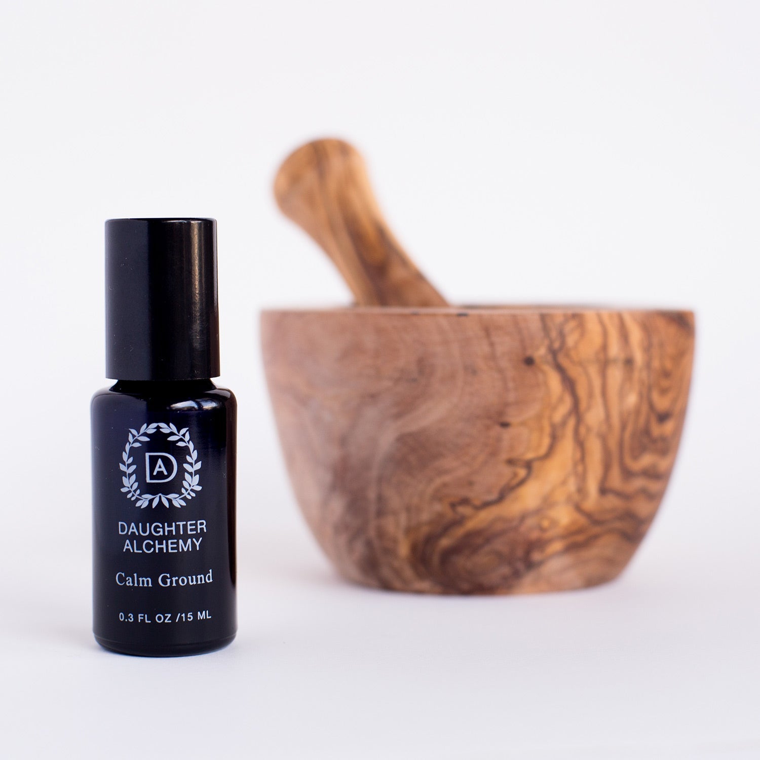 A small black bottle labeled "Calm Ground® Botanical Calming Oil" by Daughter Alchemy sits before a wooden mortar and pestle, evoking soothing organic sandalwood to calm anxiety, all set against a white background.