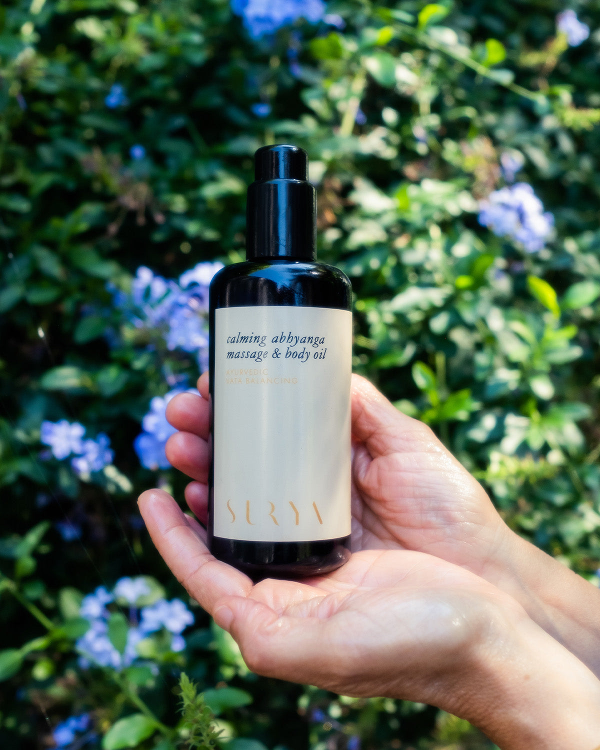A person holds a dark bottle labeled "Calming Body Oil" by Surya—ideal for soothing and balance—against a blurred backdrop of green foliage and purple flowers.