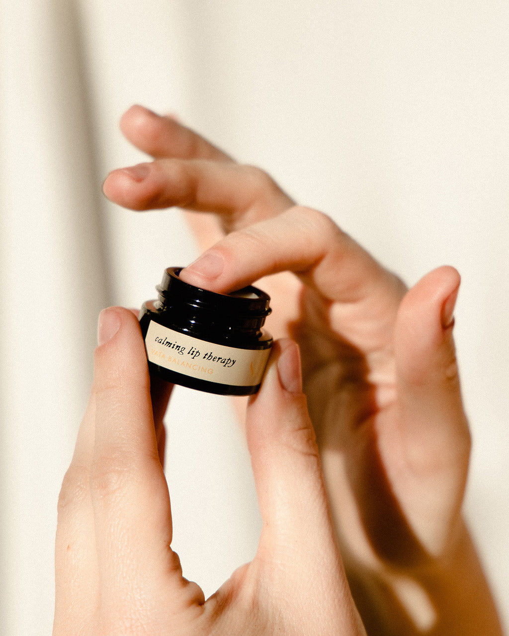 A close-up shows hands opening a Surya Calming Lip Therapy jar, with one hand holding the dark container as the other dips a finger into the soothing formula against a softly lit background.