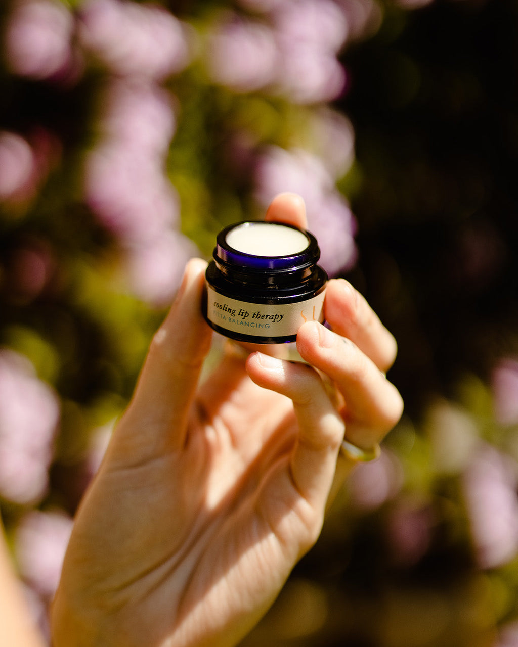 A hand holds a small, open jar of Surya Cooling Lip Therapy against a blurred background of greenery and purple flowers, highlighting its soothing and nourishing formula.