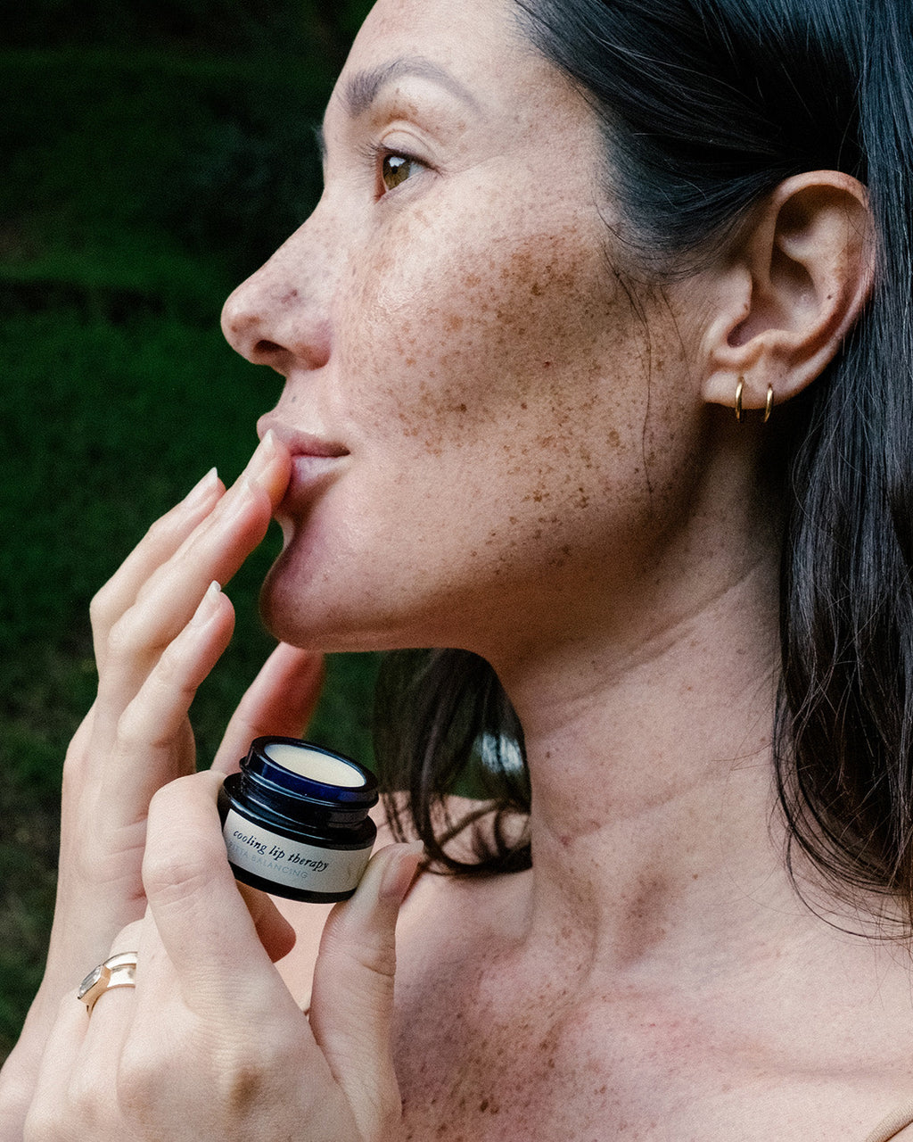 A woman with freckles applies Surya’s Cooling Lip Therapy from a small jar to her lips outdoors. She wears gold hoop earrings and rings, framed by a natural backdrop.