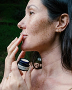 A woman with freckles applies Surya’s Cooling Lip Therapy from a small jar to her lips outdoors. She wears gold hoop earrings and rings, framed by a natural backdrop.