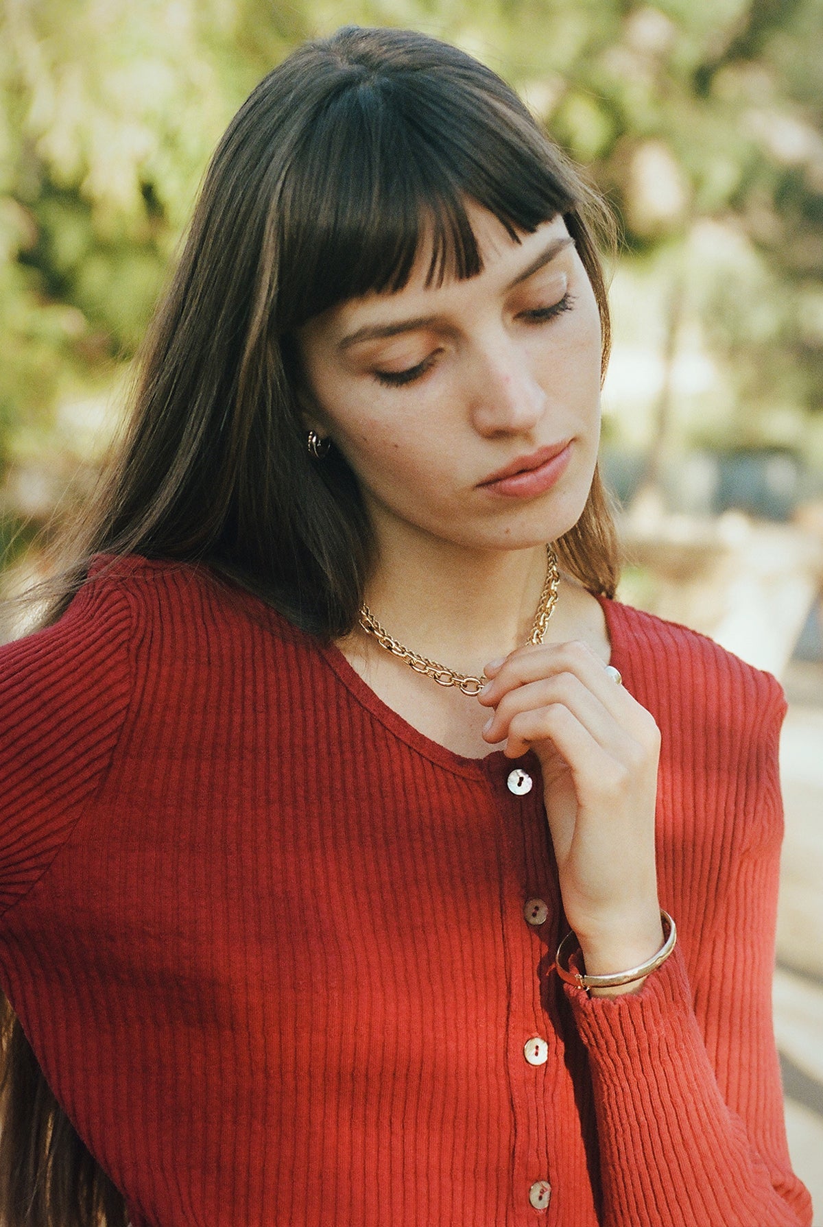 A young woman with long brown hair and bangs wears the LA RELAXED Pointelle Cardigan, featuring pearlescent buttons. She looks down thoughtfully, holding a gold necklace, against a softly blurred green outdoor background.