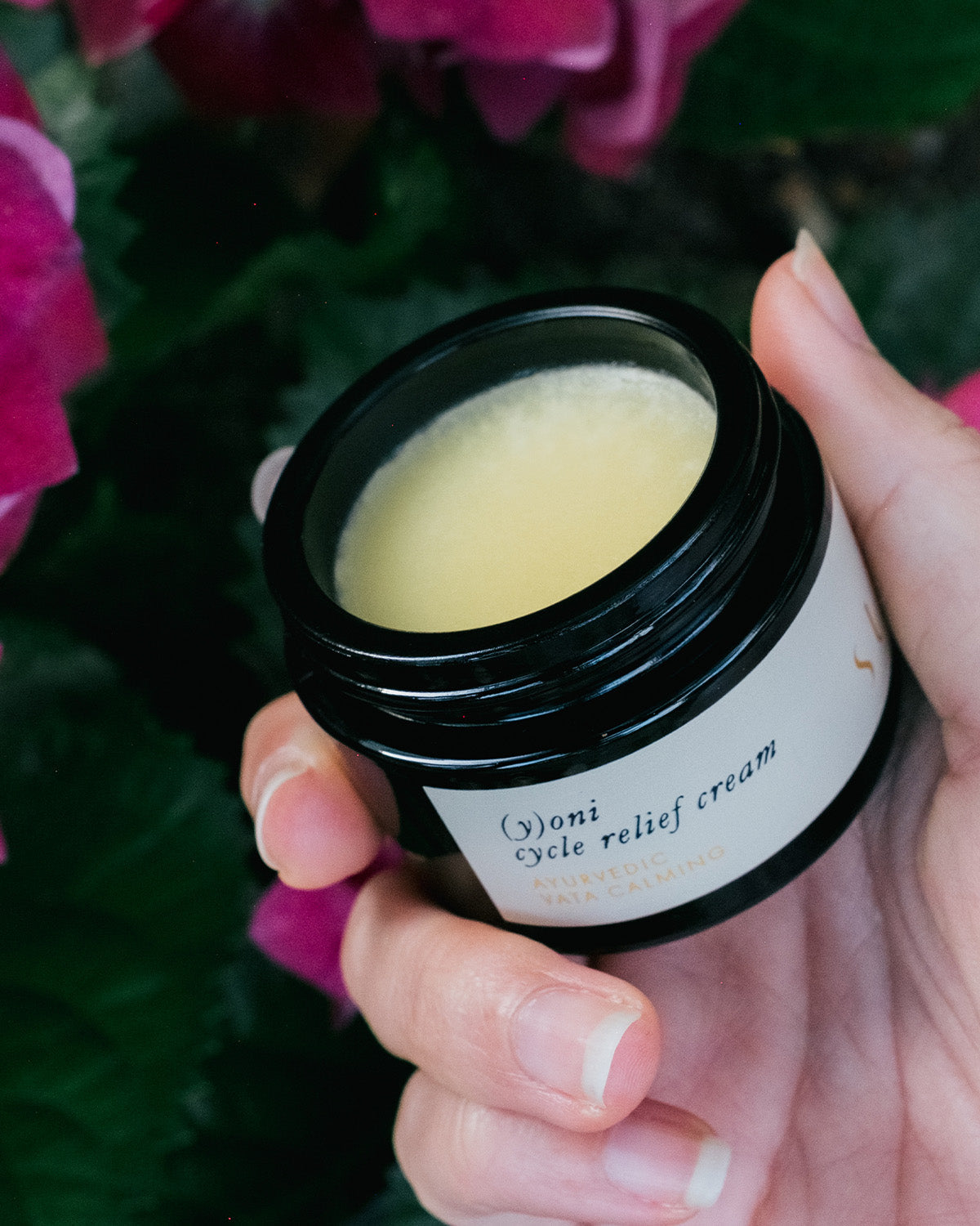 A hand holds an open jar of Surya (y)oni Cycle Relief Cream, an intimate skin moisturizer infused with Ayurvedic botanicals, with pink flowers in the background.