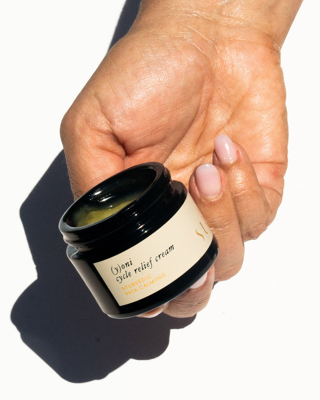 A hand holds a small, open jar of Surya (y)oni Cycle Relief Cream, an intimate moisturizer with Ayurvedic botanicals. The neatly manicured nails with pale pink polish stand out against a bright white background.