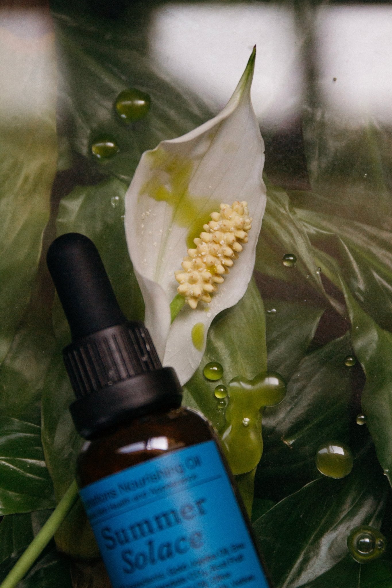 A brown glass dropper bottle labeled "Summer Solace Tallow" sits on green leaves by a white peace lily, with drops of Emanations Profound Face Oil—Deeply Nourishing Tallow, Emu Oil, Champa, Tuberose & White Ginger Lily—scattered nearby.