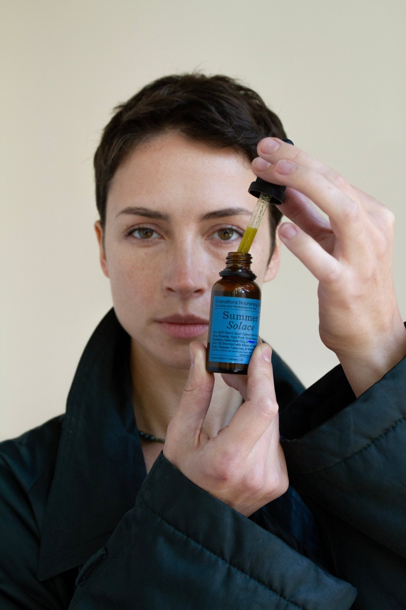 A person with short hair holds up a small brown bottle labeled "Summer Solace Tallow" and uses a dropper of Emanations Profound Face Oil—Deeply Nourishing Tallow, Emu Oil, Champa, Tuberose & White Ginger Lily—while facing the camera.