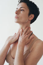 A person with short dark hair, nourished by Summer Solace Tallow’s Emanations Profound Face Oil, gently touches their neck with both hands, gazing upward serenely in a beige top against a plain light background.