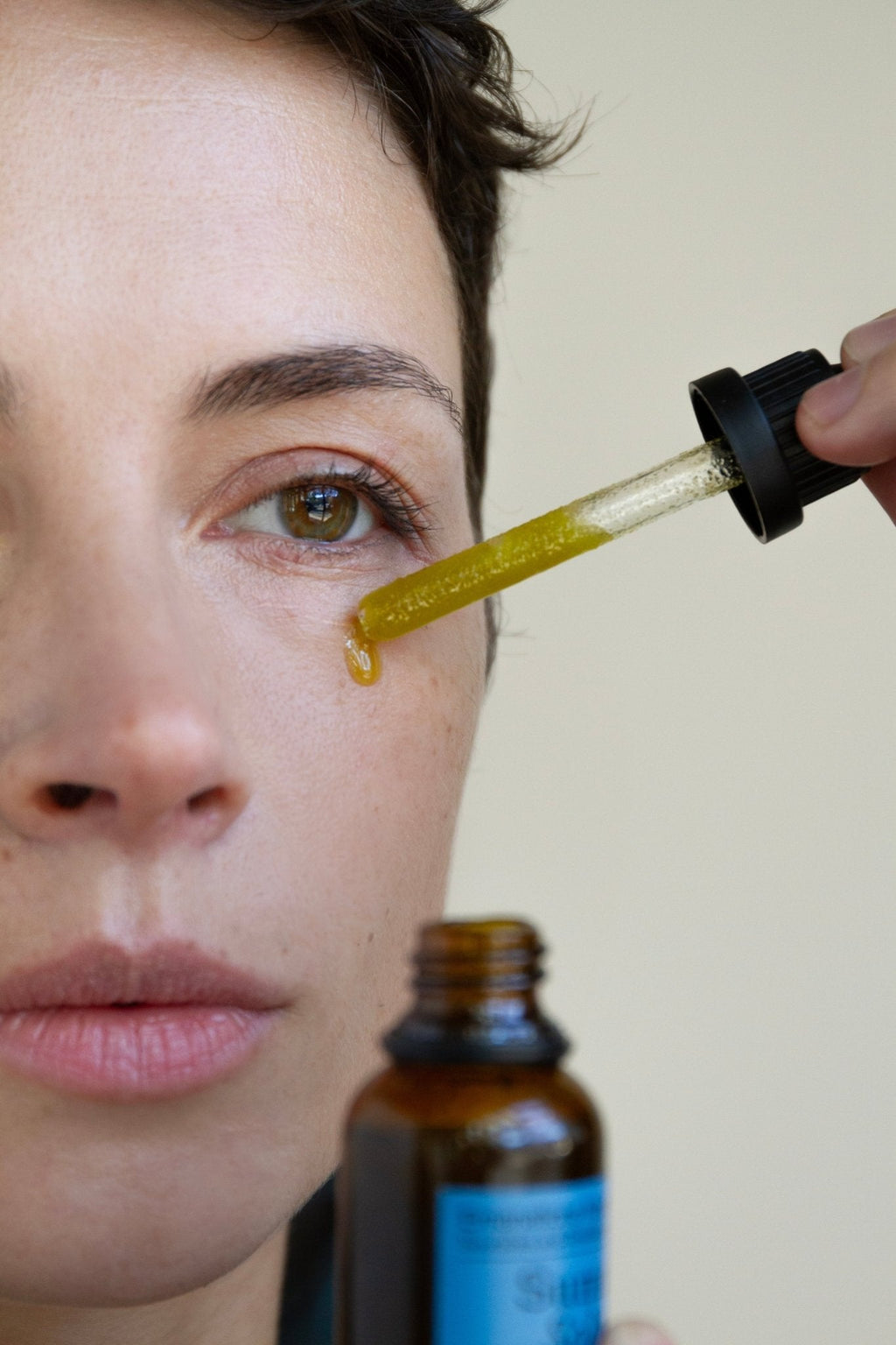 A close-up shows a person applying Summer Solace Tallow’s Emanations Profound Face Oil—featuring tallow, emu oil, champa, tuberose, and white ginger lily—under their eye with a dropper while gazing ahead with a neutral expression.