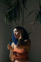 A woman with blue curly hair in a rust sleeveless top holds Summer Solace Tallow's Emanations Profound Face Oil—made with tallow, emu oil, champa, tuberose & white ginger lily—while touching her face under leafy plant shadows.