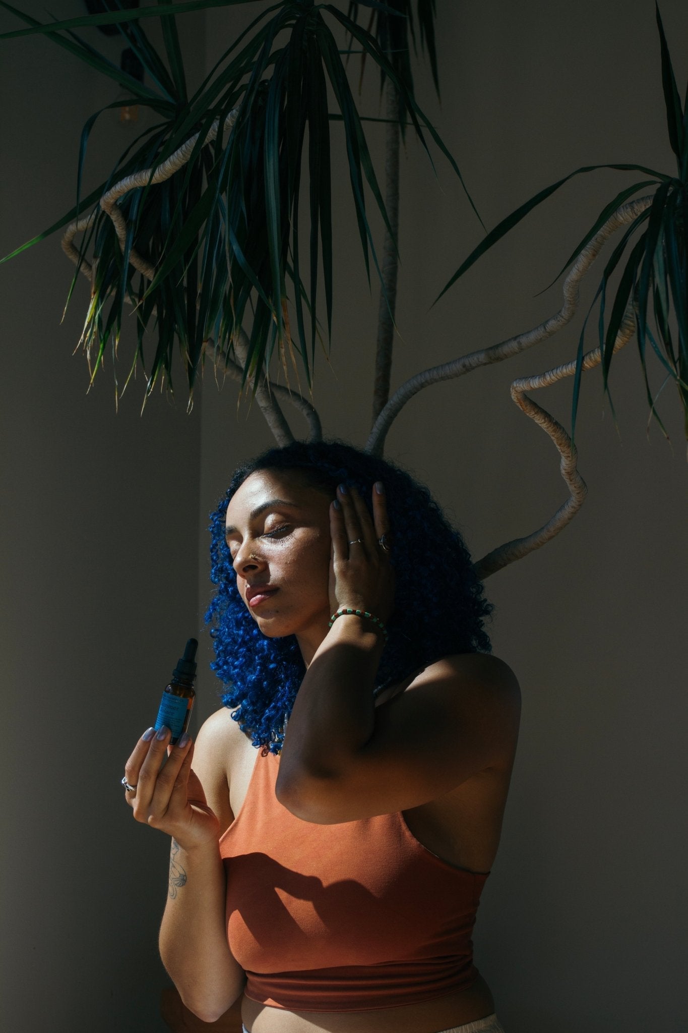 A woman with blue curly hair in a rust sleeveless top holds Summer Solace Tallow's Emanations Profound Face Oil—made with tallow, emu oil, champa, tuberose & white ginger lily—while touching her face under leafy plant shadows.