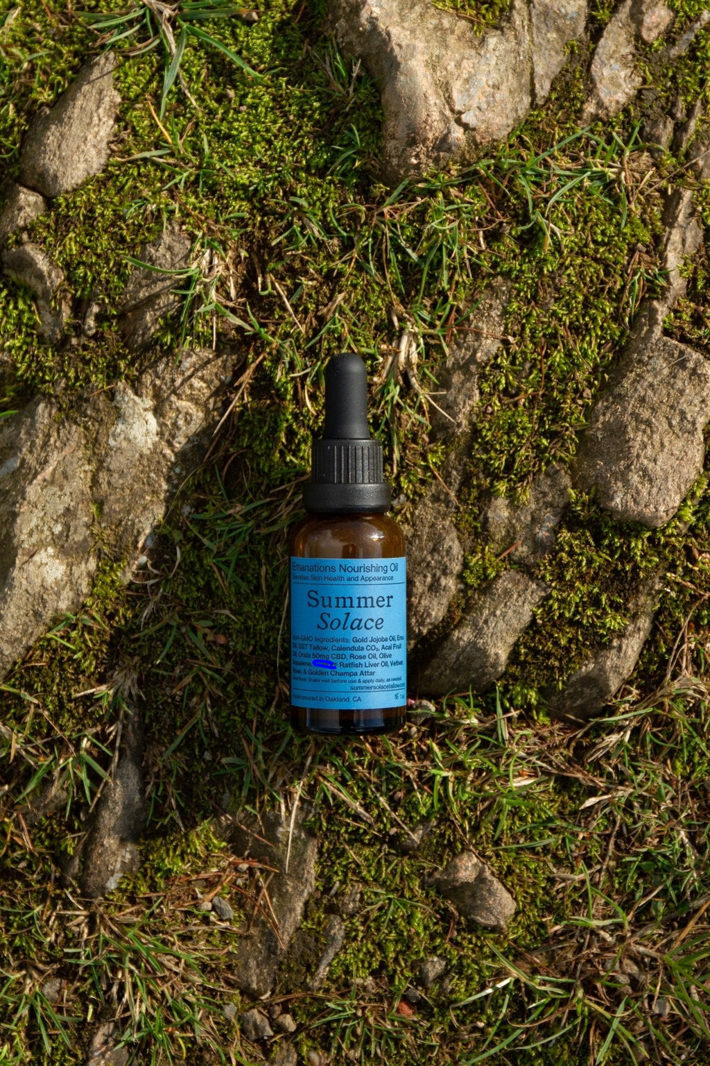 A small dark glass dropper bottle labeled "Summer Solace Tallow"—Emanations Profound Face Oil with tallow, emu oil, champa, tuberose, and white ginger lily—rests on grass and mossy rocks bathed in sunlight.