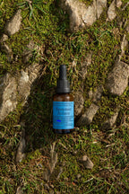 A small dark glass dropper bottle labeled "Summer Solace Tallow"—Emanations Profound Face Oil with tallow, emu oil, champa, tuberose, and white ginger lily—rests on grass and mossy rocks bathed in sunlight.