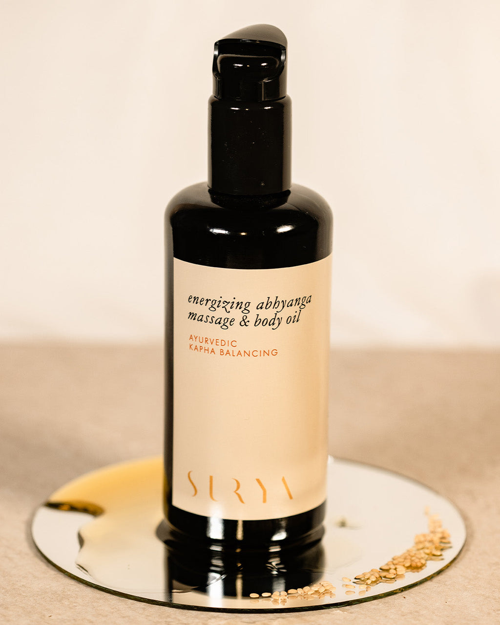 A black bottle of Surya Energizing Body Oil rests on a small round mirror with scattered sesame seeds, featuring a beige label with black and gold text, ideal for balancing Kapha dosha, set against a neutral backdrop.