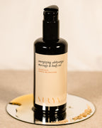 A black bottle of Surya Energizing Body Oil rests on a small round mirror with scattered sesame seeds, featuring a beige label with black and gold text, ideal for balancing Kapha dosha, set against a neutral backdrop.