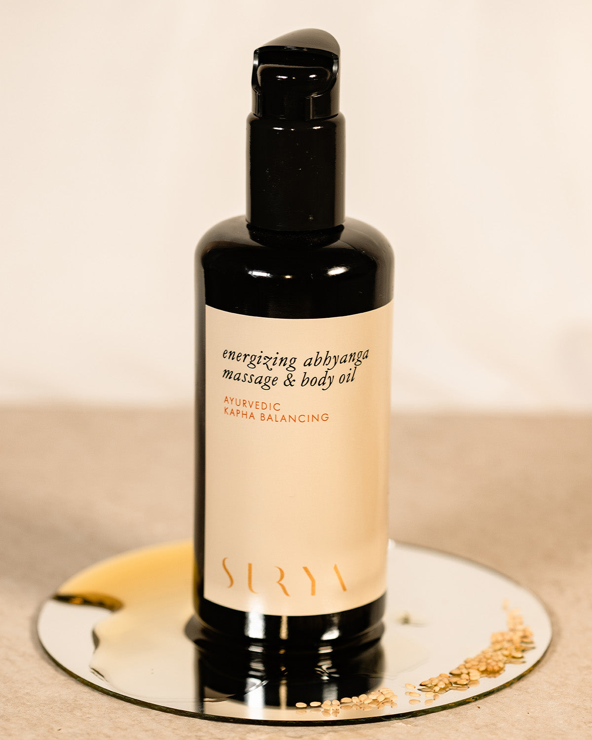 A black bottle of Surya Energizing Body Oil rests on a small round mirror with scattered sesame seeds, featuring a beige label with black and gold text, ideal for balancing Kapha dosha, set against a neutral backdrop.