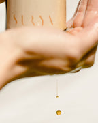 A hand pours Surya Energizing Body Oil from a beige bottle into another open palm, with drops of oil caught midair against a white background.