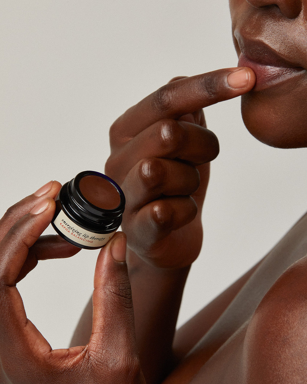 A person applies Surya Energizing Lip Therapy to their lips with a finger, holding a small jar. The neutral background emphasizes the lips and hands, highlighting this natural Lip Therapy moment.