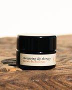 A small dark jar of Surya Energizing Lip Therapy rests on a wooden surface with a light background, offering nourishing care through its natural formula.