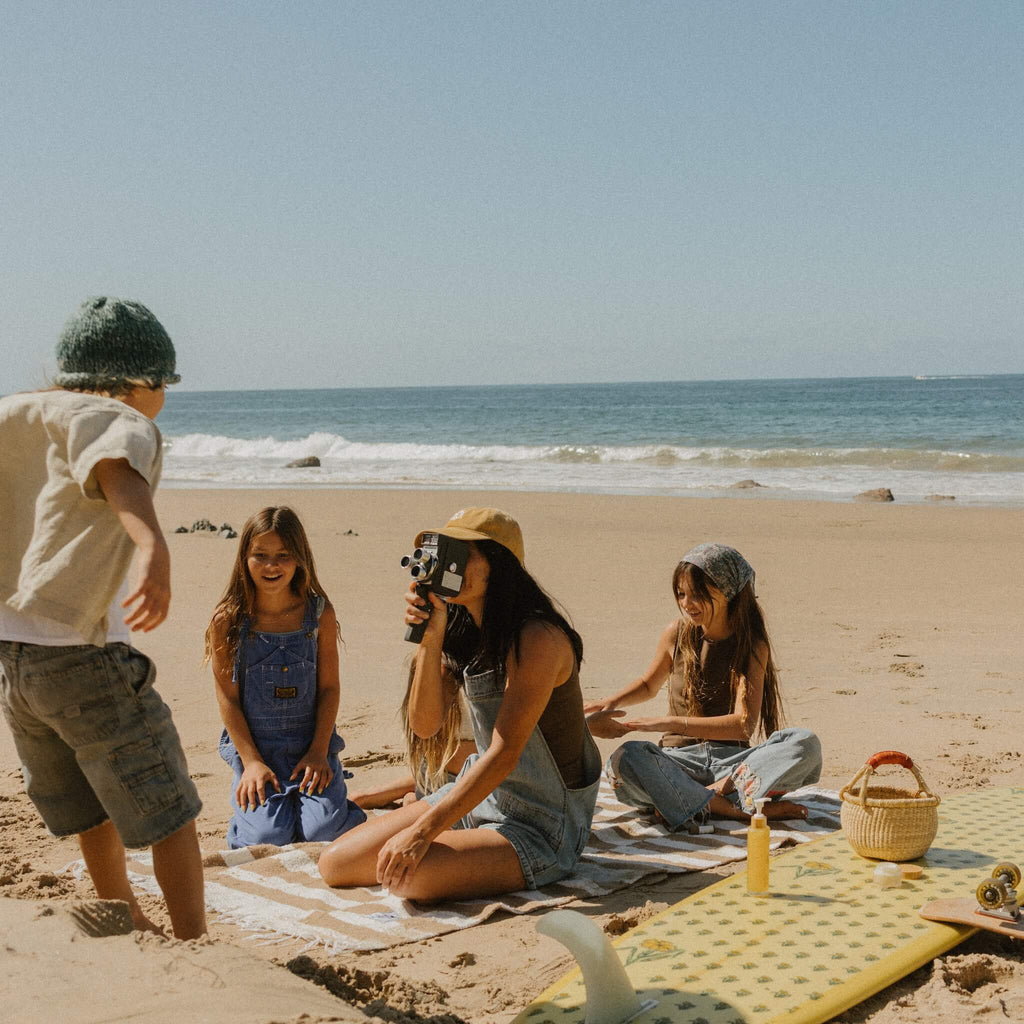 A woman with glowing skin enjoys a beach day with three kids, a surfboard, and a picnic by the ocean, capturing the moment on camera. She radiates California Sunshine by Honeybee Hippie.