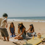 A woman with glowing skin enjoys a beach day with three kids, a surfboard, and a picnic by the ocean, capturing the moment on camera. She radiates California Sunshine by Honeybee Hippie.
