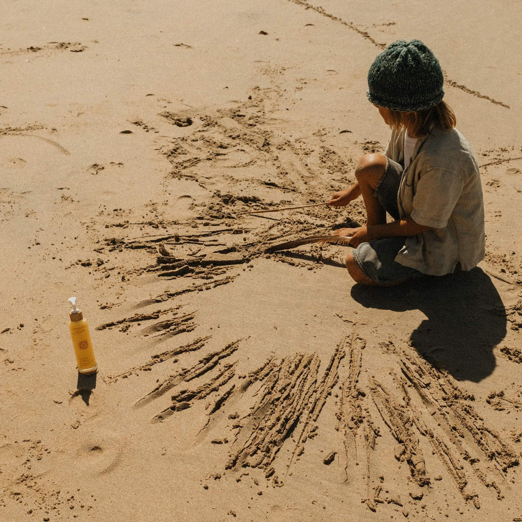 A child in a green knit hat draws in the sand with sticks, sitting cross-legged near a bottle of Honeybee Hippie’s california sunshine body oil, their glowing skin radiant under the warm beach sun.