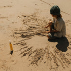 A child in a green knit hat draws in the sand with sticks, sitting cross-legged near a bottle of Honeybee Hippie’s california sunshine body oil, their glowing skin radiant under the warm beach sun.