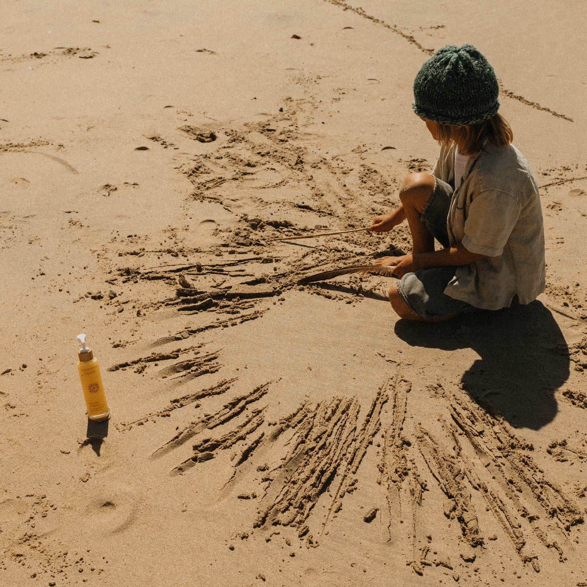 A child in a green knit hat draws in the sand with sticks, sitting cross-legged near a bottle of Honeybee Hippie’s california sunshine body oil, their glowing skin radiant under the warm beach sun.