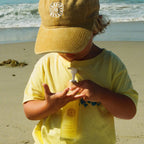 A young child in a yellow shirt and cap stands on the beach, applying Honeybee Hippie’s california sunshine body oil for glowing skin, as waves and sand shimmer gently in the background.