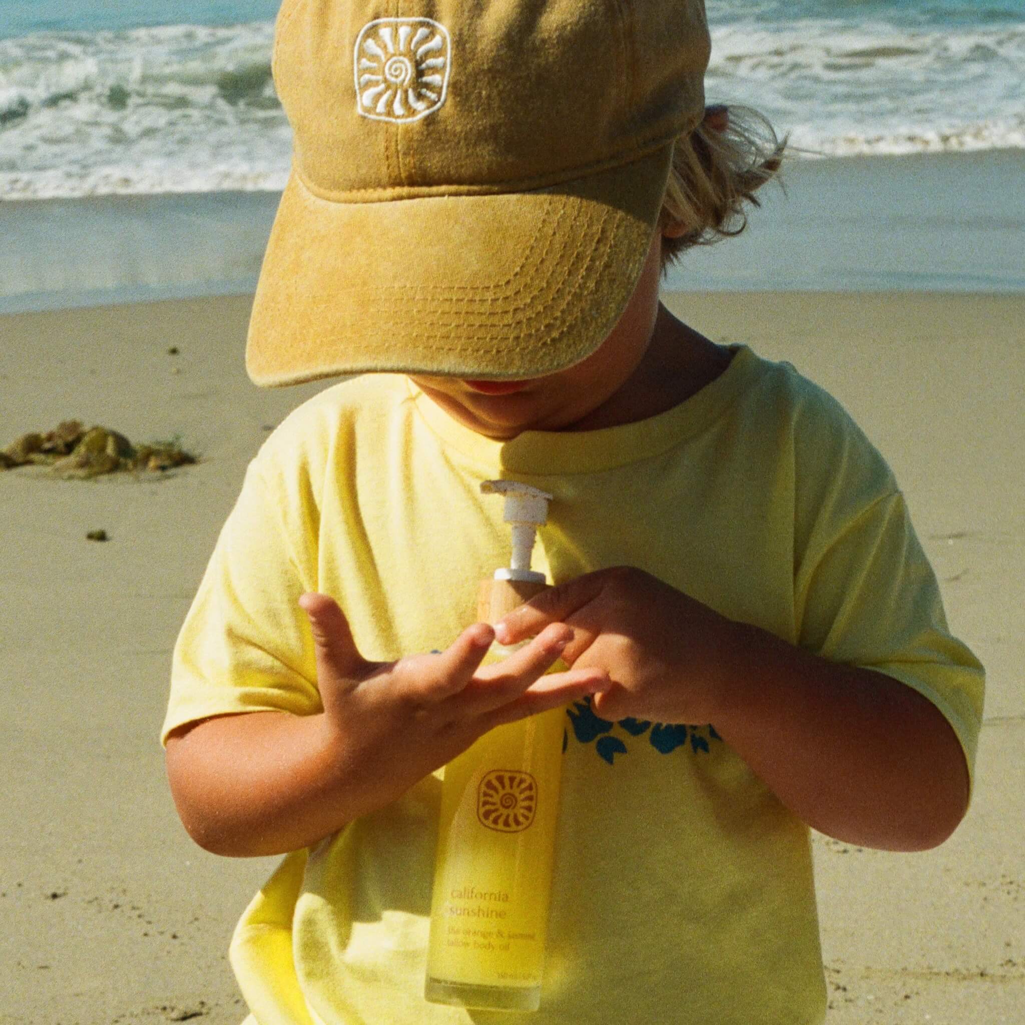 A young child in a yellow shirt and cap stands on the beach, applying Honeybee Hippie’s california sunshine body oil for glowing skin, as waves and sand shimmer gently in the background.