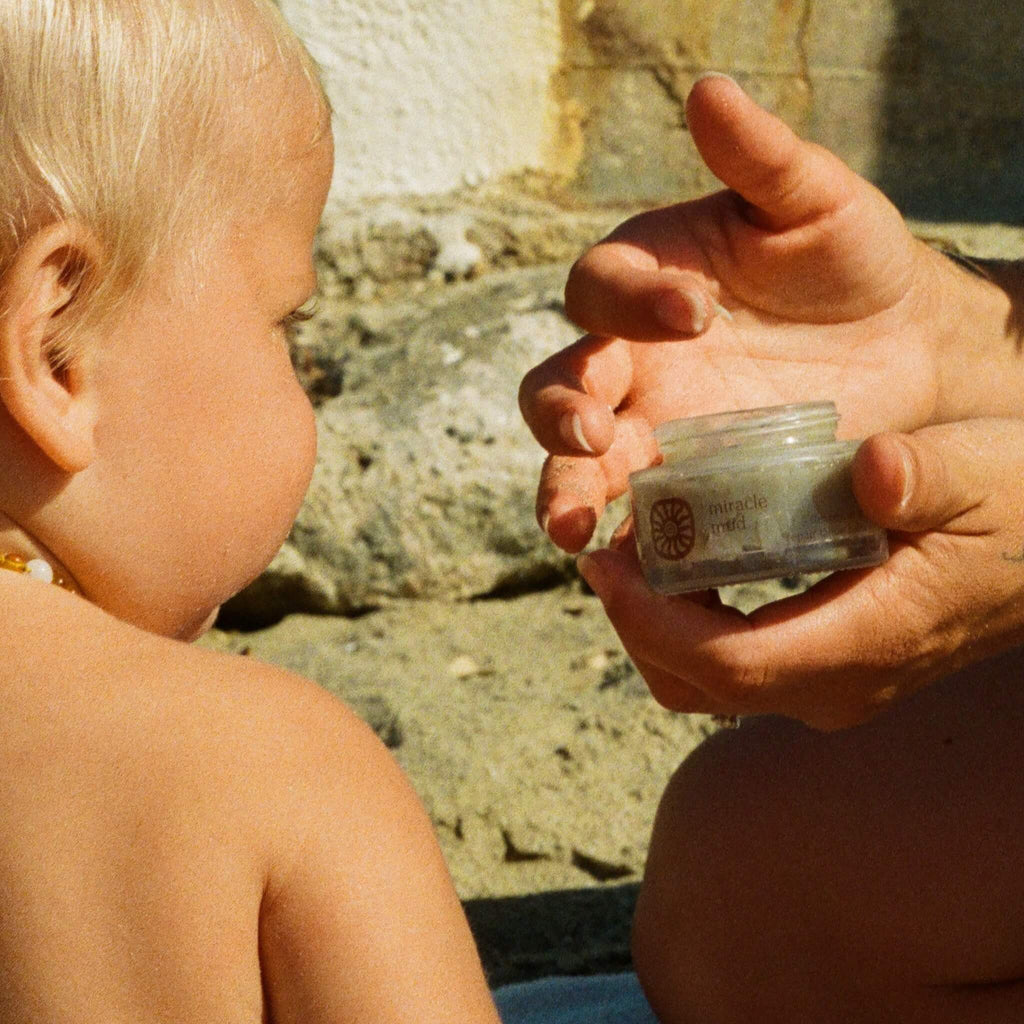 A young child observes an adult holding an open jar of Honeybee Hippie's miracle mud outdoors, with sand and a stone wall as the backdrop.