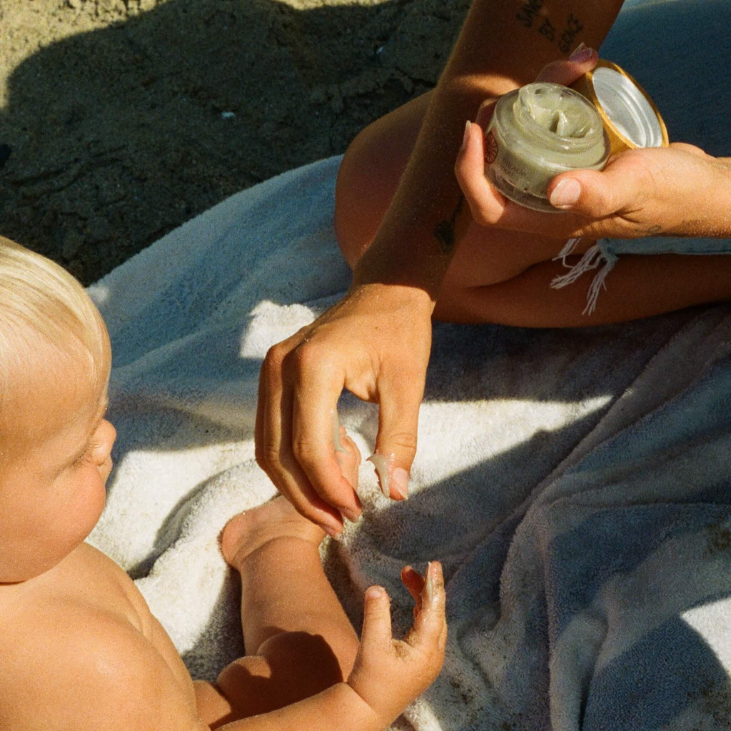 An adult gently rubs Honeybee Hippie’s miracle mud onto a light-haired baby’s arm while they sit on a beach towel, providing soothing care under the sun.