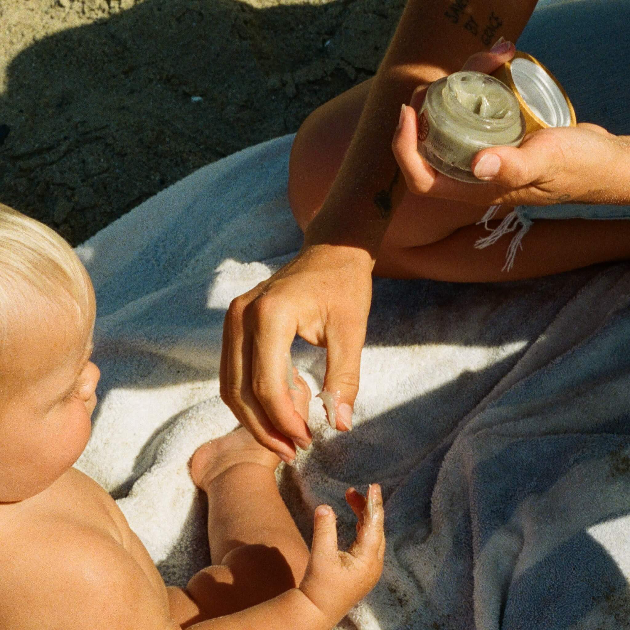 An adult gently rubs Honeybee Hippie’s miracle mud onto a light-haired baby’s arm while they sit on a beach towel, providing soothing care under the sun.