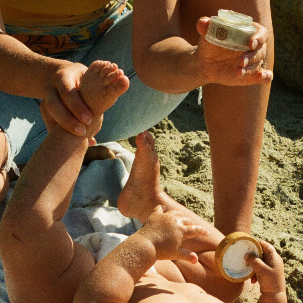 A baby relaxes on the beach, holding a container as an adult gently applies Honeybee Hippie’s miracle mud tallow cream from a jar, highlighting a tender moment of care and protection.