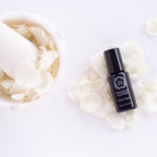 A black bottle labeled "Heart Salve® Botanical Nurturing Oil" by Daughter Alchemy rests on white rose petals beside a mortar and pestle, emphasizing its calming blend with organic white rose oil against a pristine white background.