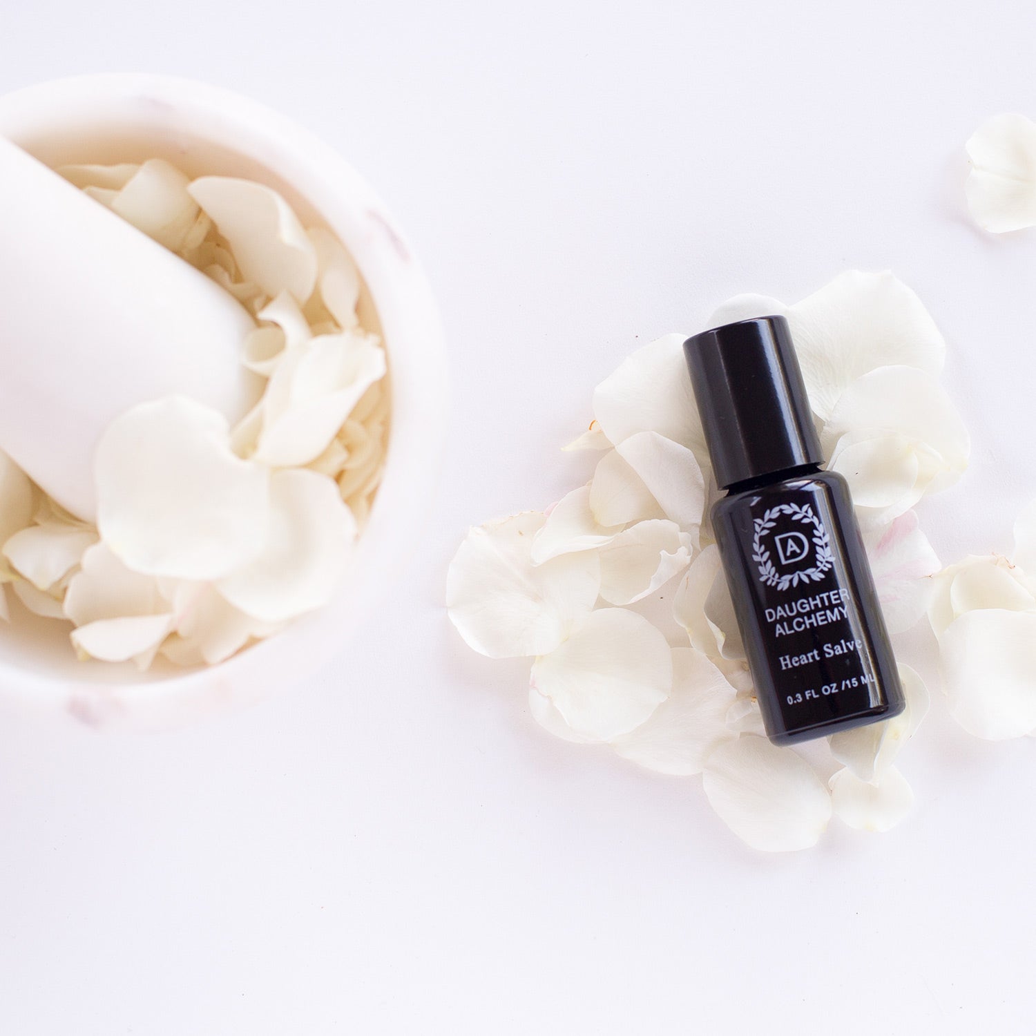 A black bottle labeled "Heart Salve® Botanical Nurturing Oil" by Daughter Alchemy rests on white rose petals beside a mortar and pestle, emphasizing its calming blend with organic white rose oil against a pristine white background.