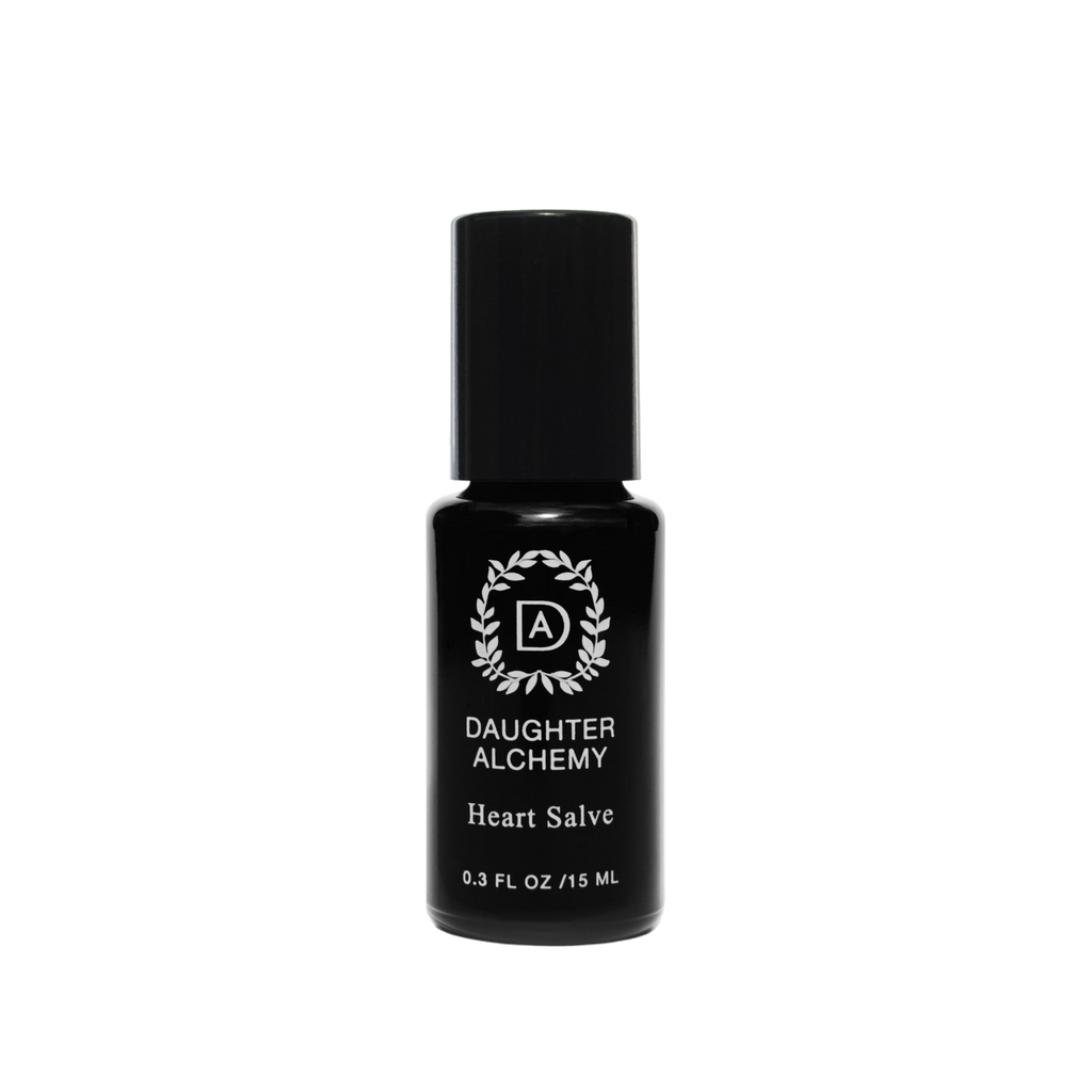 A small black bottle of Heart Salve® Botanical Nurturing Oil by Daughter Alchemy, crafted with organic ingredients, is displayed against a black background. The label features a white logo with a wreath design around the initials "DA" above the product name and indicates the bottle contains 0.3 fl oz (15 ml) of nurturing oil salve.