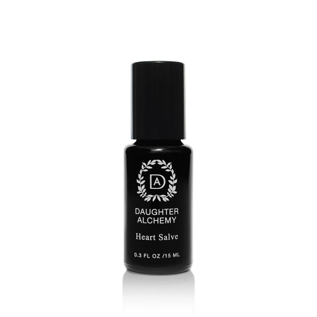 A black bottle with a black cap, labeled "Daughter Alchemy Heart Salve® Botanical Nurturing Oil" and "0.3 FL OZ / 15 ML" in white text, features a circular leaf logo above the brand name; it blends organic white rose oil for soothing comfort.