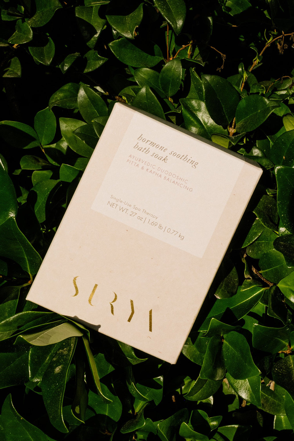 A cream-colored box labeled "Hormone Soothing Bath Soak" by Surya, 27 oz (1.68 lb/0.77 kg), rests on lush green leaves, with product details shown on the packaging.