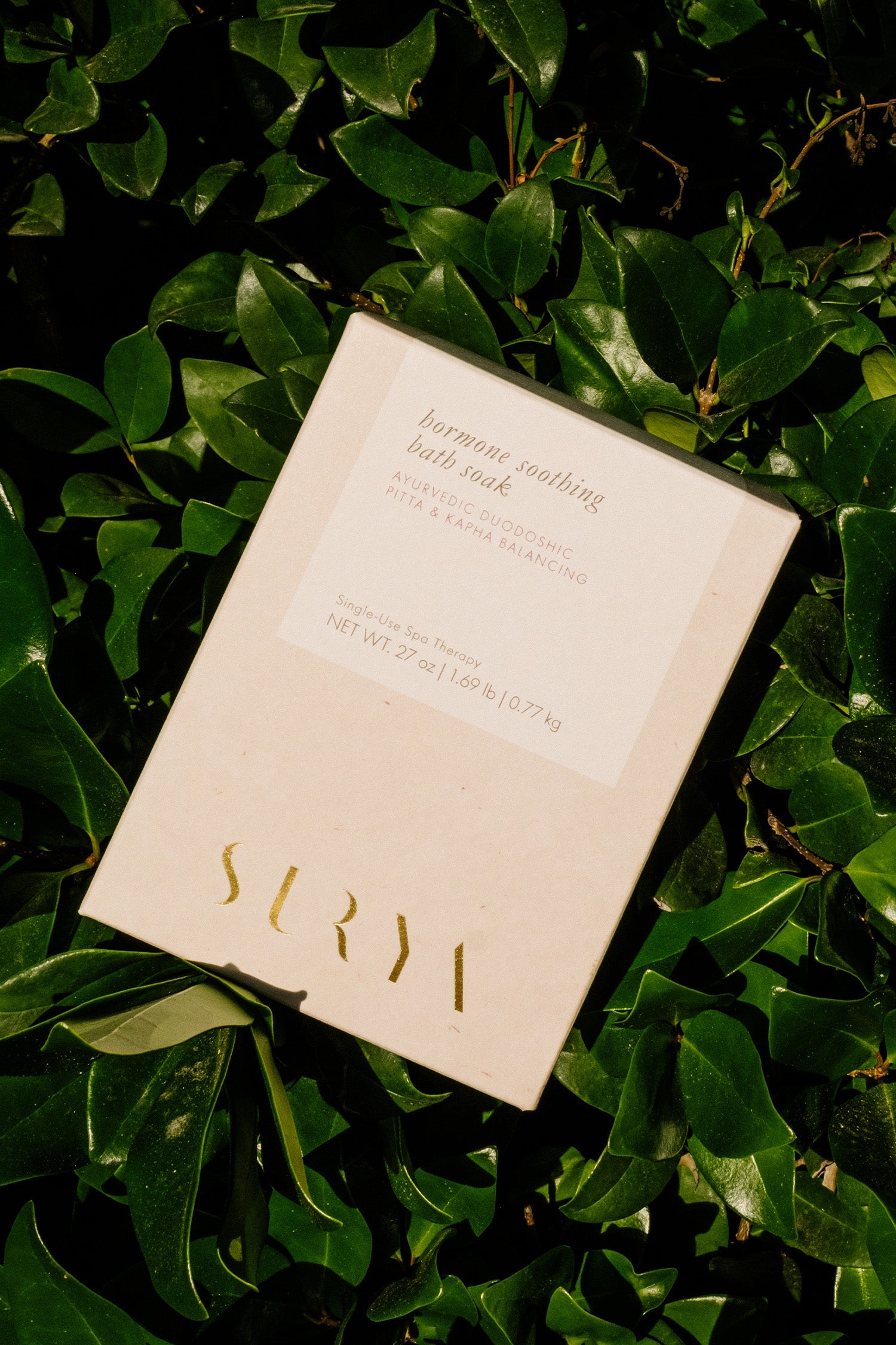 A cream-colored box labeled "Hormone Soothing Bath Soak" by Surya, 27 oz (1.68 lb/0.77 kg), rests on lush green leaves, with product details shown on the packaging.