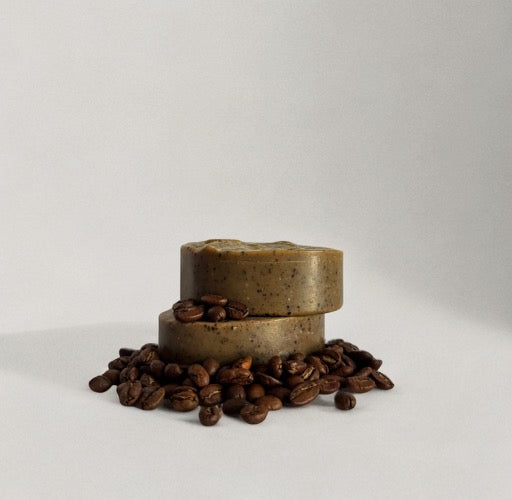 Two round bars of Wild Mamas Shilajit + Decaffeinated Coffee Bean Deeply Exfoliating Wild Bison Tallow Soap Scrub stacked together with scattered coffee beans—ideal exfoliation for sensitive skin using only natural ingredients.