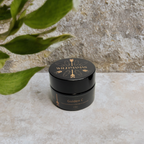 A small black jar labeled "Wild Mamas Golden C - Wild Bison Tallow Balm" rests on a light stone surface with a leafy plant in the foreground and textured stone background, highlighting this skincare balm.