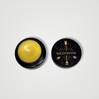 A small round black container of Golden C - Wild Bison Tallow Balm with yellow balm inside sits open. The lid, beside the container, displays the Wild Mamas logo with gold arrows and feather designs on black.