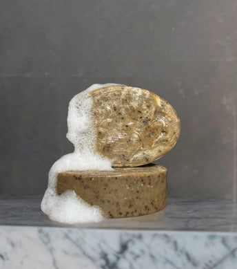 Two natural Wild Mamas soap bars, one oval and one round, stacked on dark gray marble with foamy suds. Featuring Shilajit + Decaffeinated Coffee Bean- Deeply Exfoliating Wild Bison Tallow Soap Scrub for sensitive skin.