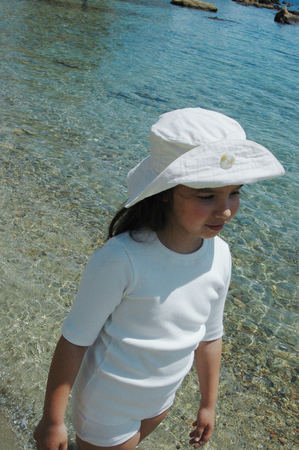 A young child dressed in ethically handmade summer wear and a "Friends of the Sun" hat by cabane childrenswear is standing at the edge of a clear, shallow body of water. The child looks out toward the distance with the sun shining, and the water appears calm with rocks visible in the background.
