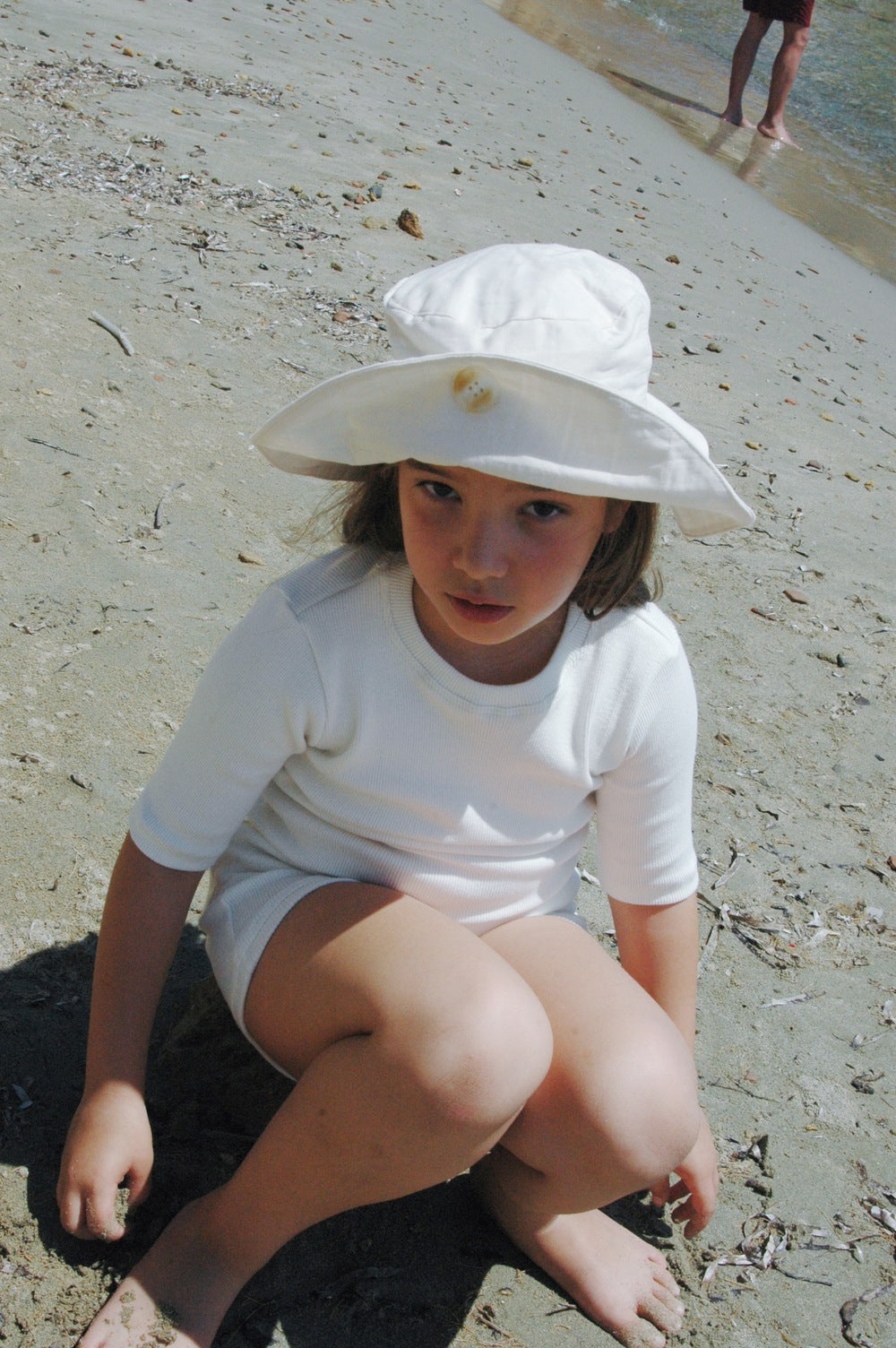 A young child wearing a "Friends of the Sun" Hat from cabane childrenswear and dressed in white summer attire sits on a sandy beach. The child looks towards the camera with the shoreline and another person visible in the background. The child appears to be in a shady area with a mix of sunlight and shadow.