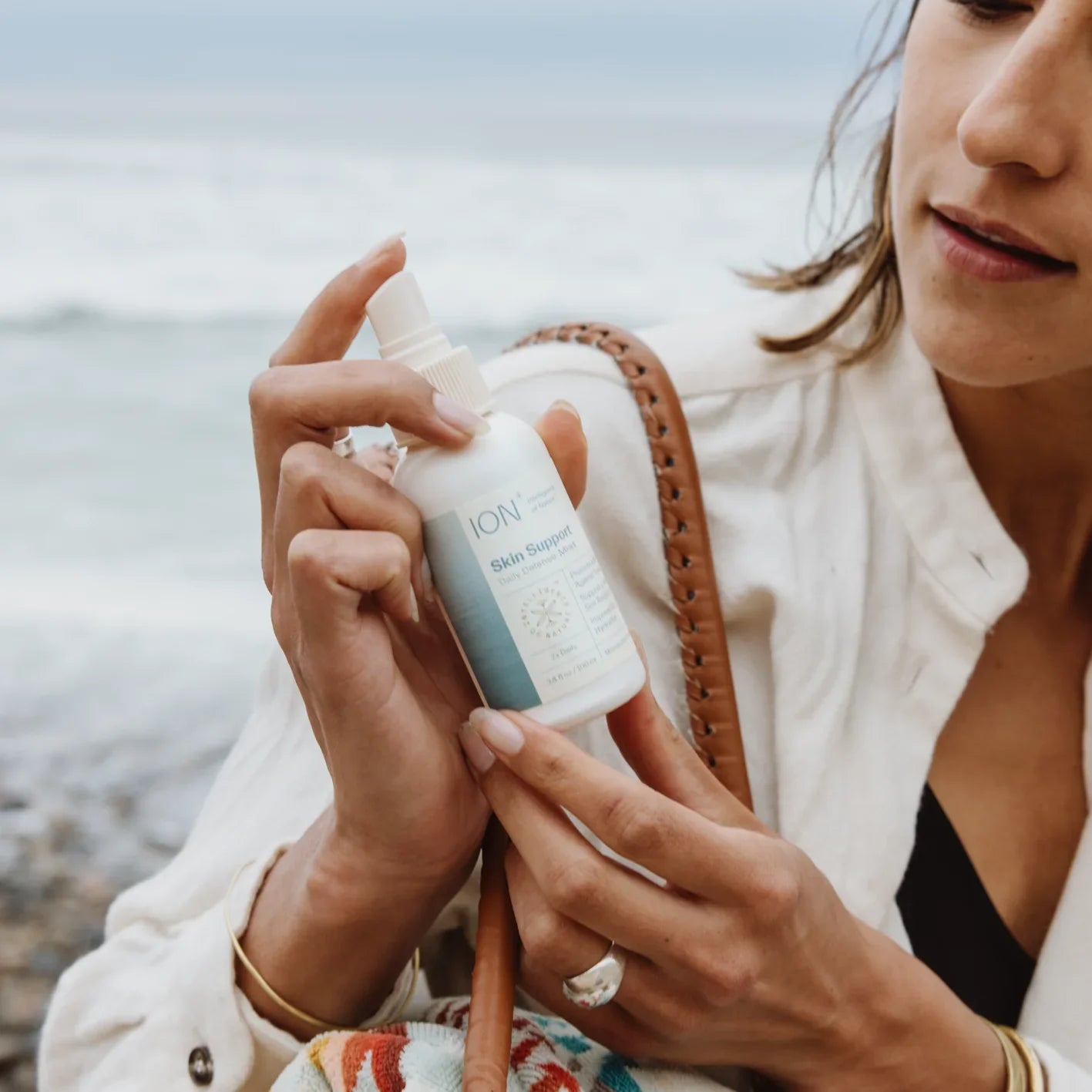 A person is holding a bottle of ION* Skin Support by the beach, eyeing the white label with blue and grey text. Clad in a white jacket and carrying a woven bag on their shoulder, they admire it as waves crash in the background—perfect for maintaining your skin's microbiome barrier.