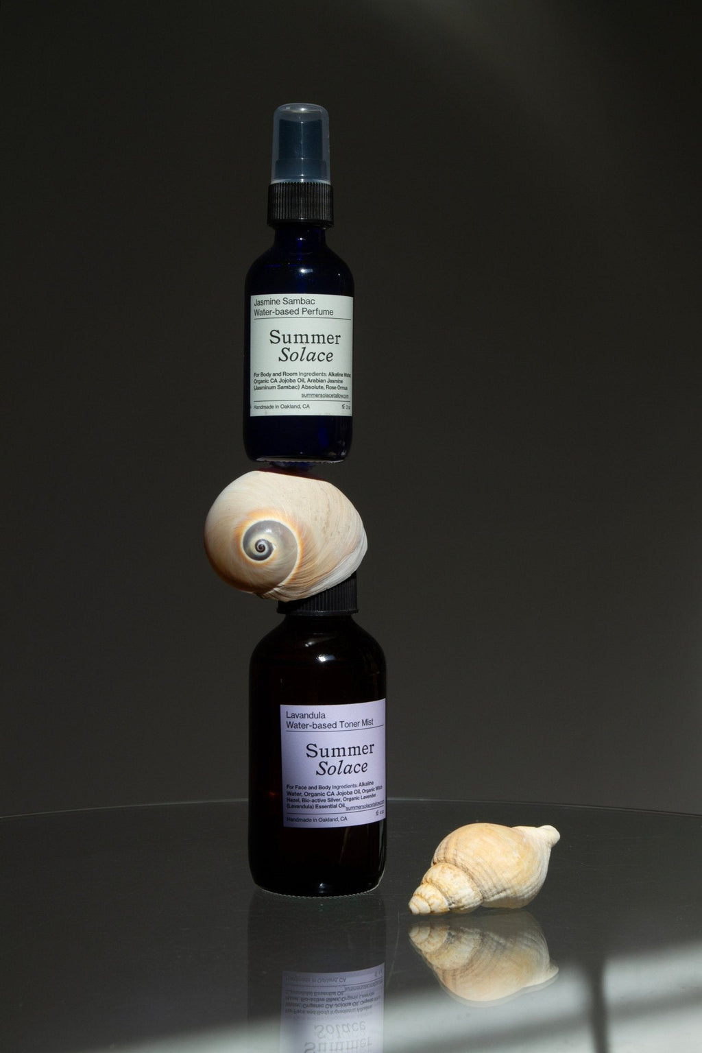 Two dark glass bottles of Summer Solace Tallow's Jasmine Sambac + Magnolia Perfume Mist—Water-Based are stacked with a seashell between them; another seashell sits nearby on a reflective surface against a plain, dark background.