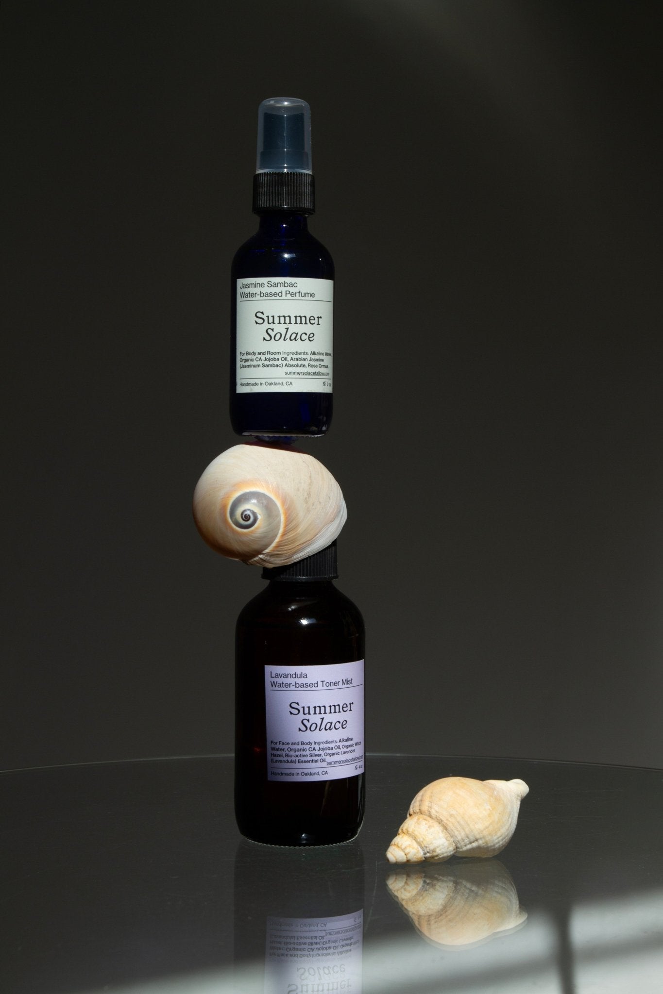 Two dark glass bottles of Summer Solace Tallow's Jasmine Sambac + Magnolia Perfume Mist—Water-Based are stacked with a seashell between them; another seashell sits nearby on a reflective surface against a plain, dark background.