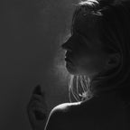 A woman in profile sprays Summer Solace Tallow's Jasmine Sambac + Magnolia Perfume Mist – Water-Based onto her face, with soft lighting highlighting the mist’s droplets and casting dramatic shadows across her features.
